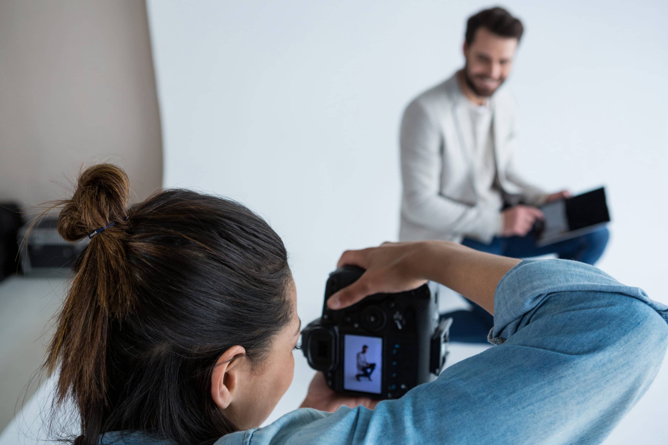 Kvinnlig fotograf tittar i kameran under fotografering av en man som sitter en bit bort i någon form av studiomiljö