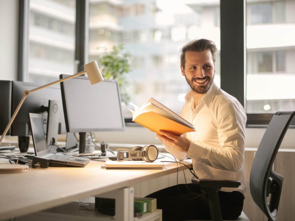 En glad person håller i en bok och sitter vid en skrivbordsdator i kontorsmiljö